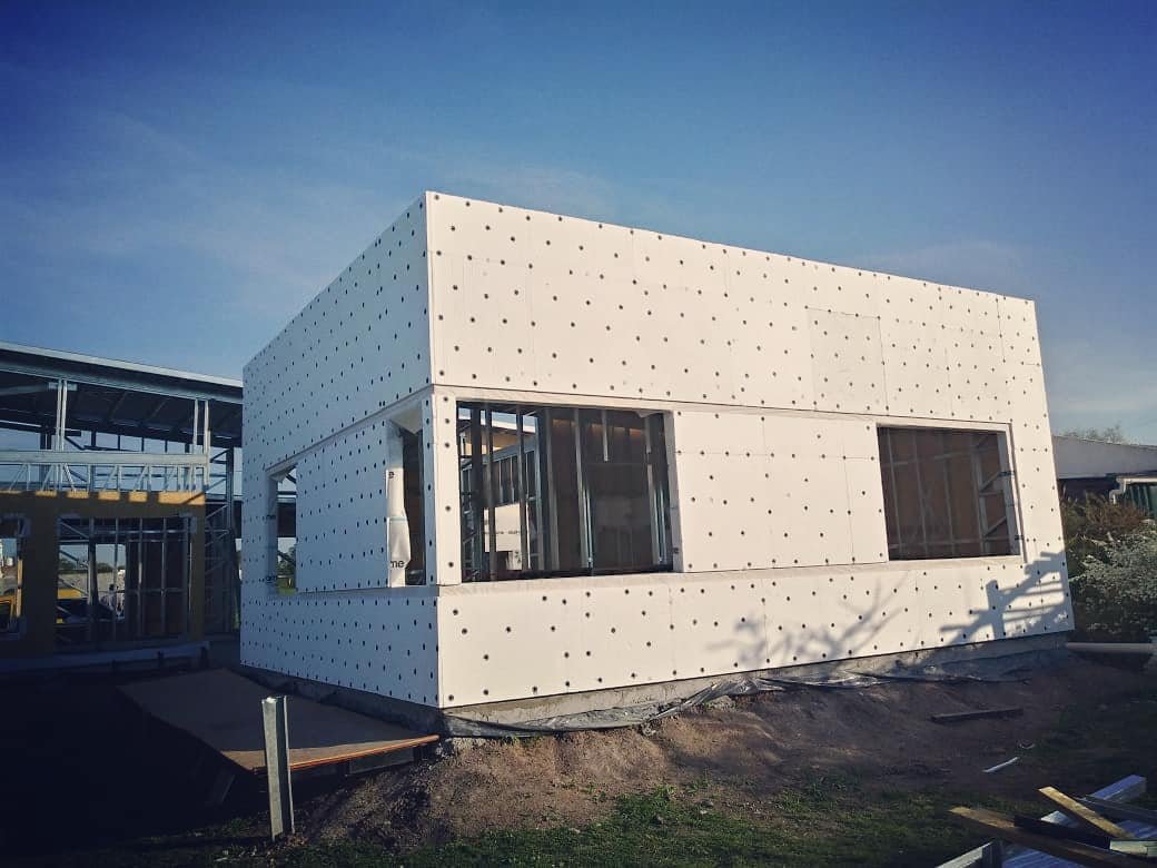 Vista de uma construção em Light Steel Frame com painéis de isolamento branco instalados em toda a fachada. A estrutura metálica interna fica visível pelas janelas abertas; o terreno ao redor é de terra batida e o céu está limpo.